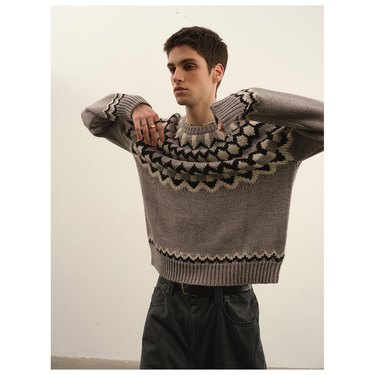 Model wearing a taupe gray sweater with a black and cream geometric yoke and wide dark trousers, arms raised.