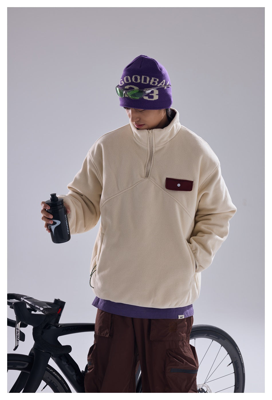 Model in cream half-zip jacket holding a water bottle beside a road bicycle, wearing beanie and brown pants
