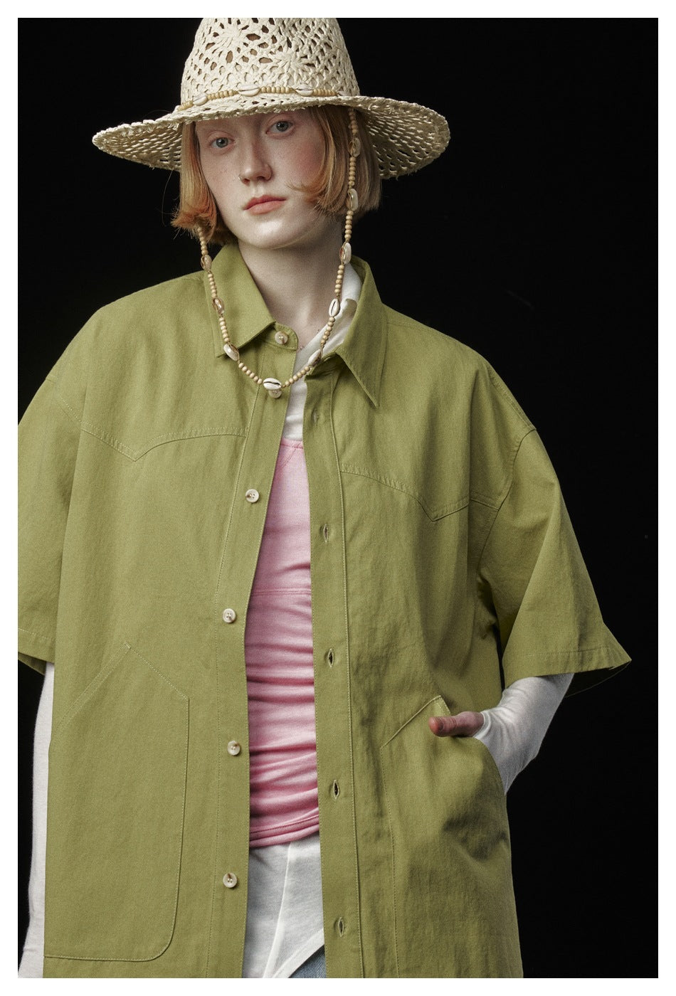 Close view of an olive-green shirt with large patch pockets, worn over a pink top by a model in a woven hat.