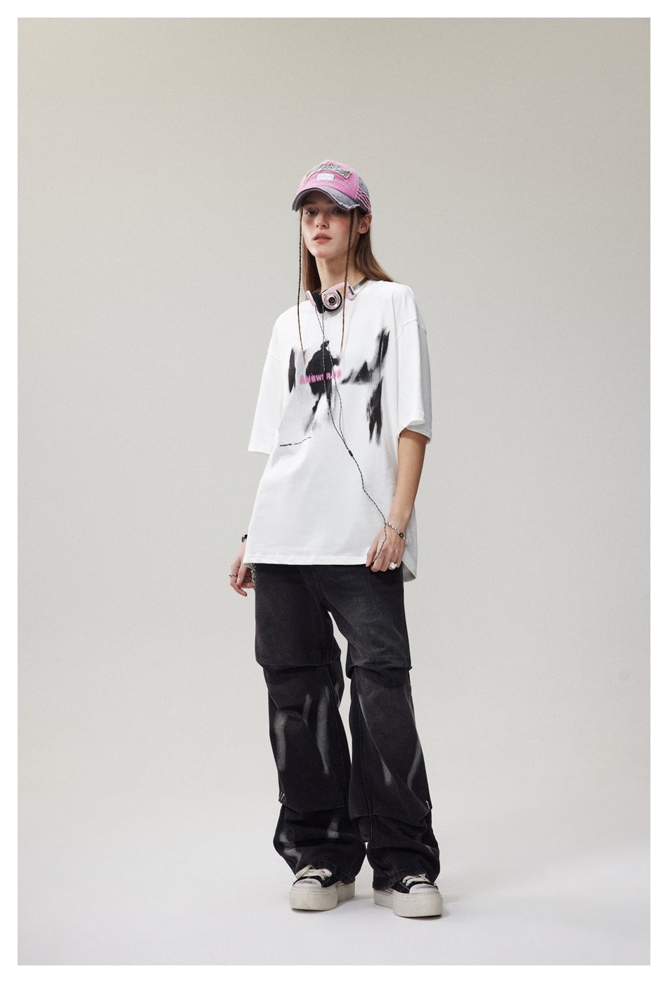 Angled view of a model in an oversized white T-shirt with black and pink chest artwork and loose black trousers.