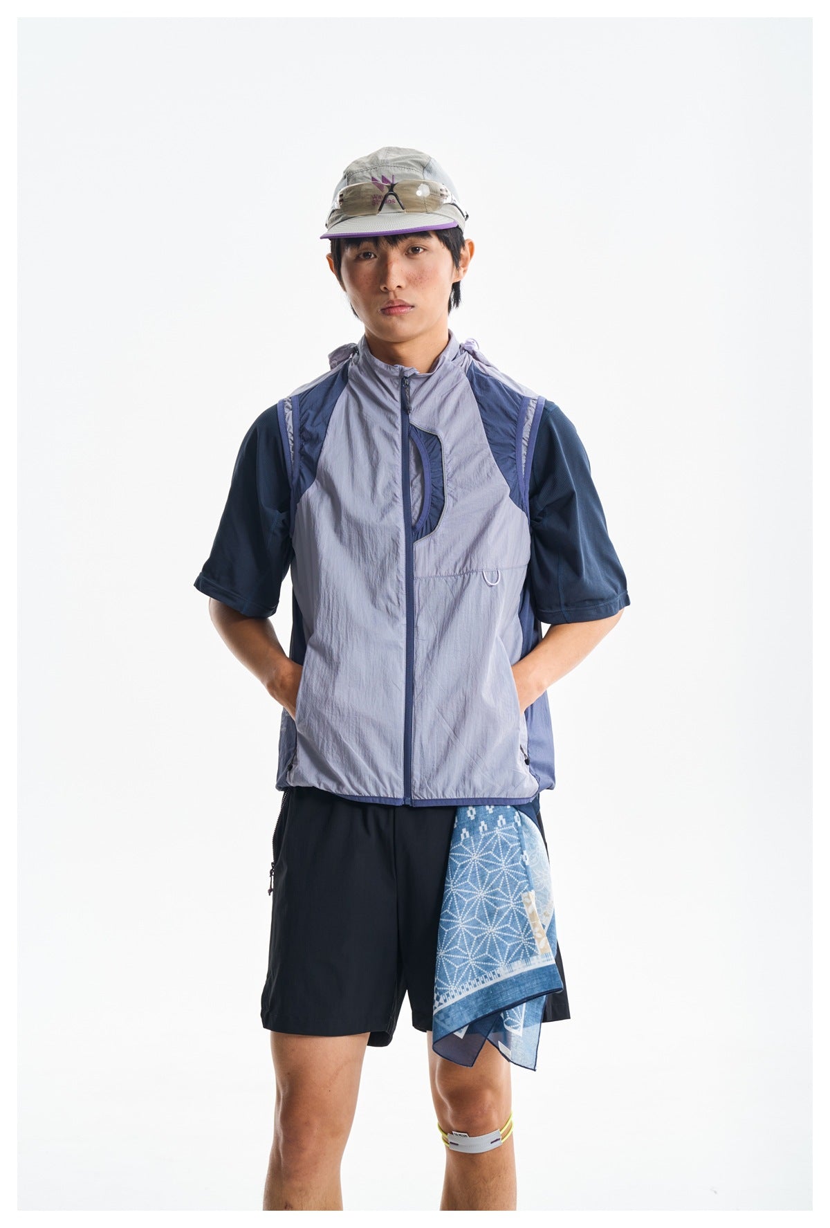 Model wearing a light lavender zip-front vest over a navy shirt, black shorts, and a blue patterned cloth at the hip.
