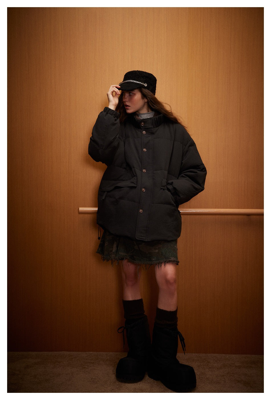 Model wearing a black quilted jacket with a high collar, front snaps and large pockets, paired with a patterned skirt.