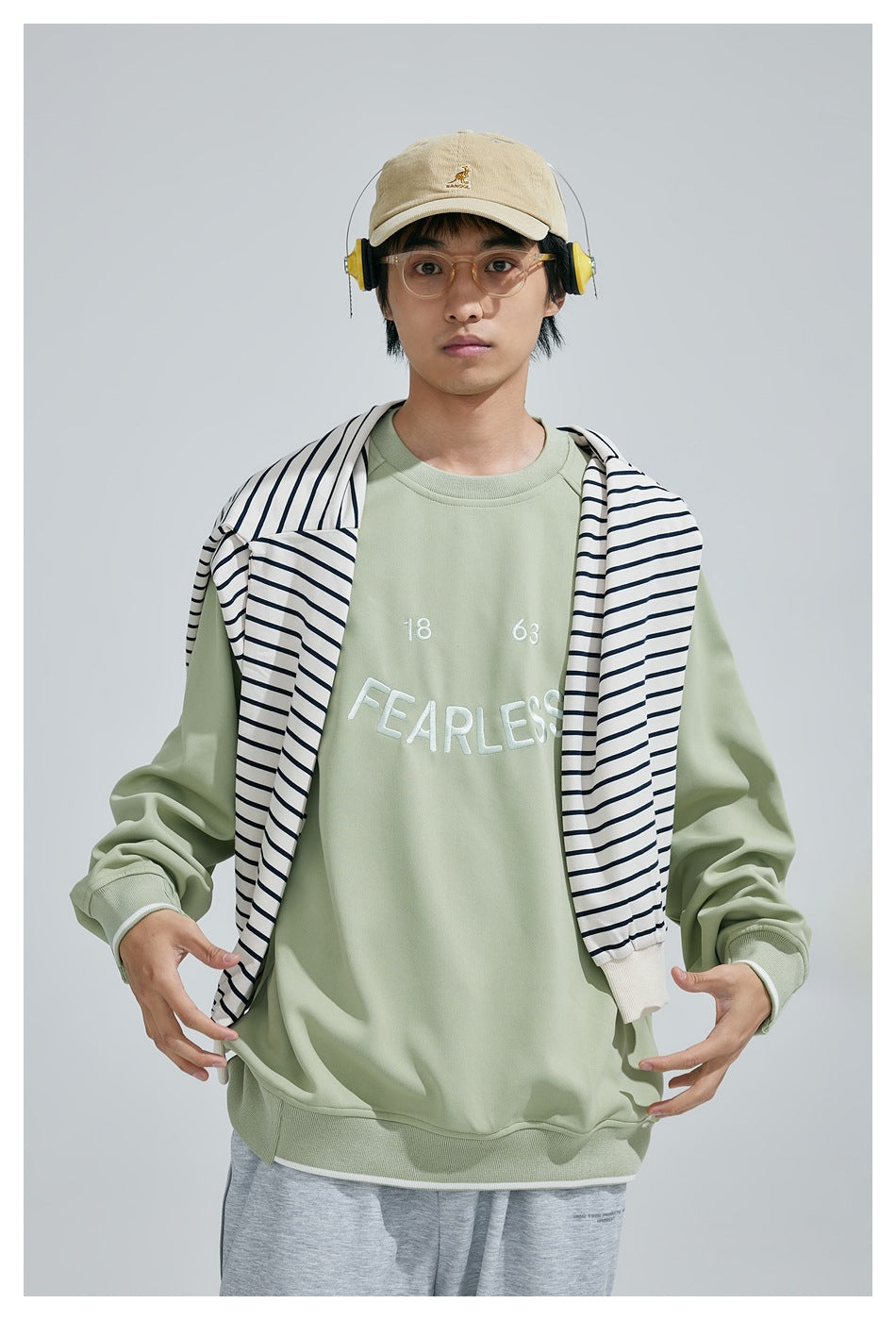 Model wearing a sage-green sweatshirt embroidered with “18 63 FEARLESS,” with a navy-striped garment draped over his
