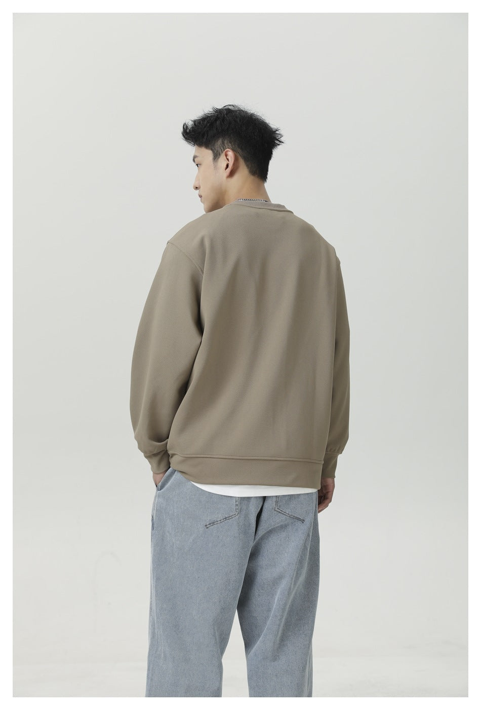 Rear three-quarter view of a man wearing a taupe crewneck sweatshirt and light blue jeans.