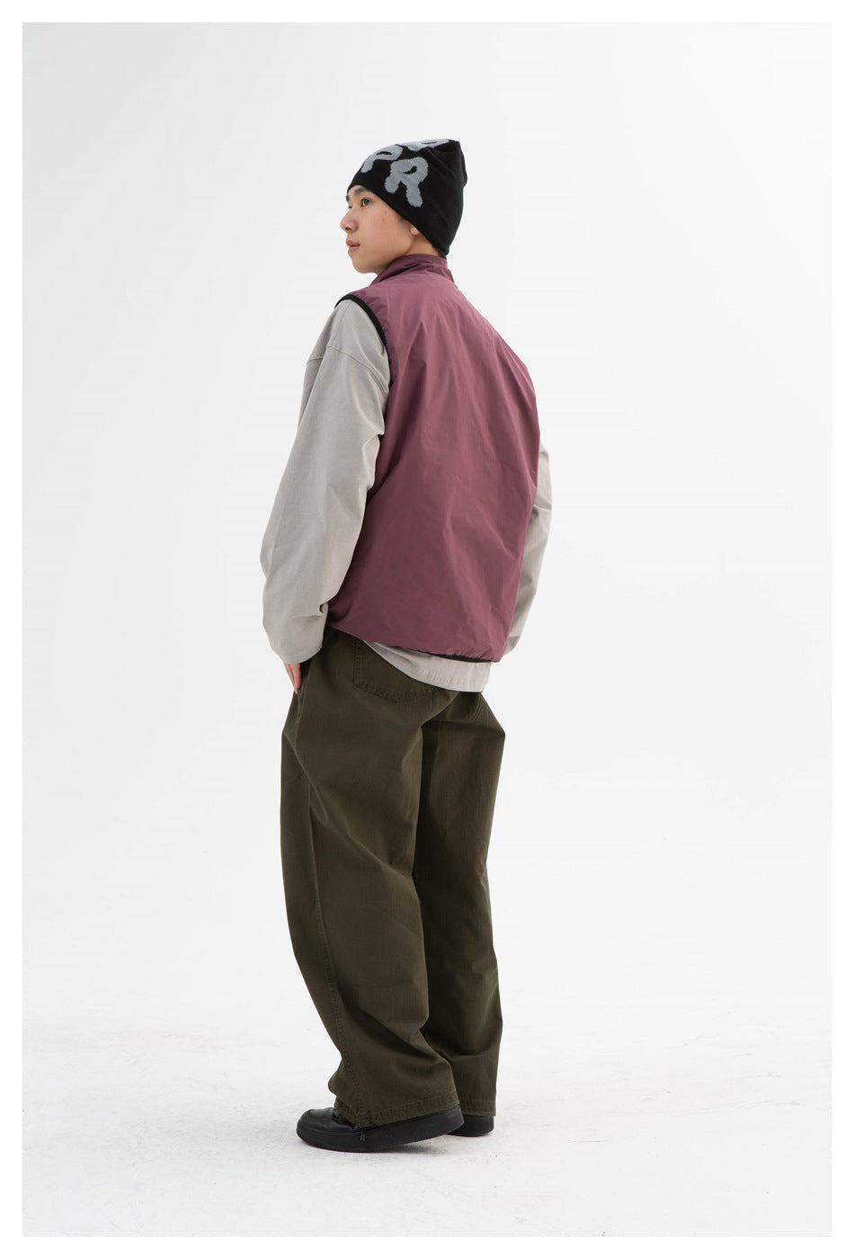 Model wearing a plum vest over a gray long-sleeve top and olive trousers, shown in side-back view