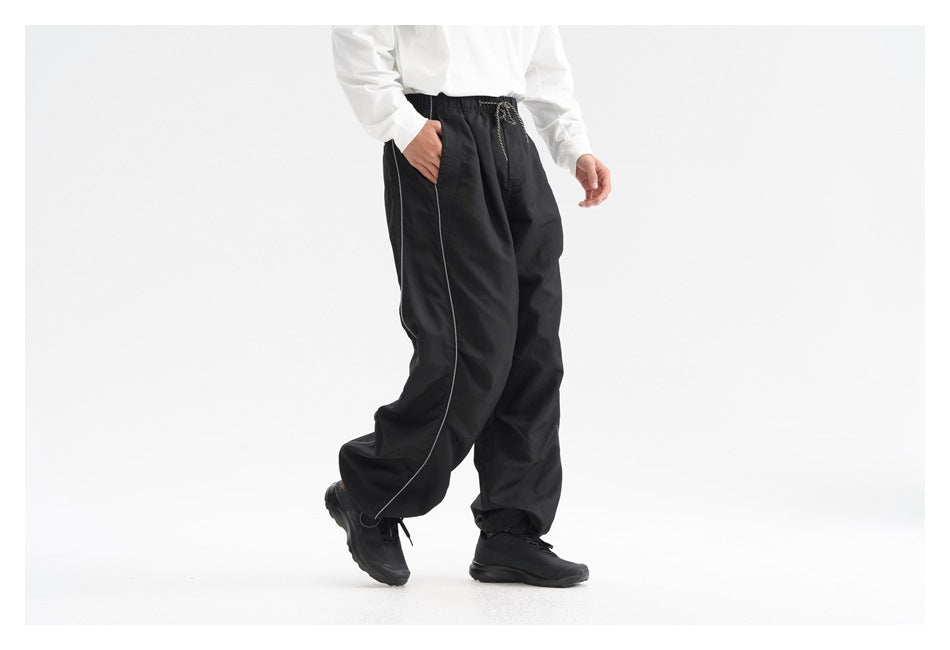Cropped model wearing black relaxed track pants with an elastic drawstring waist, light piping and gathered ankles.