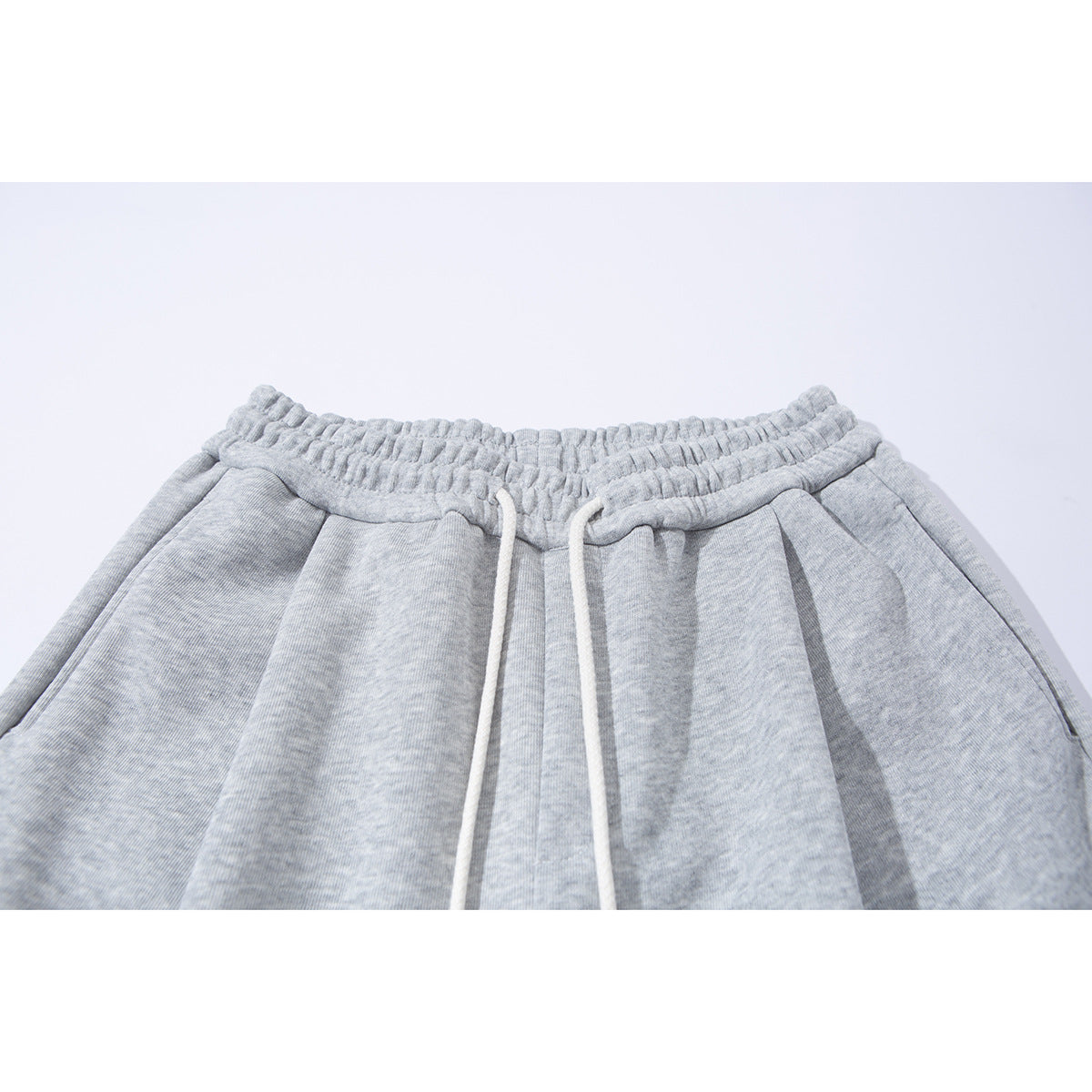 Close-up of light gray sweatpants showing the gathered elastic waistband, white drawstrings, and front pleats.