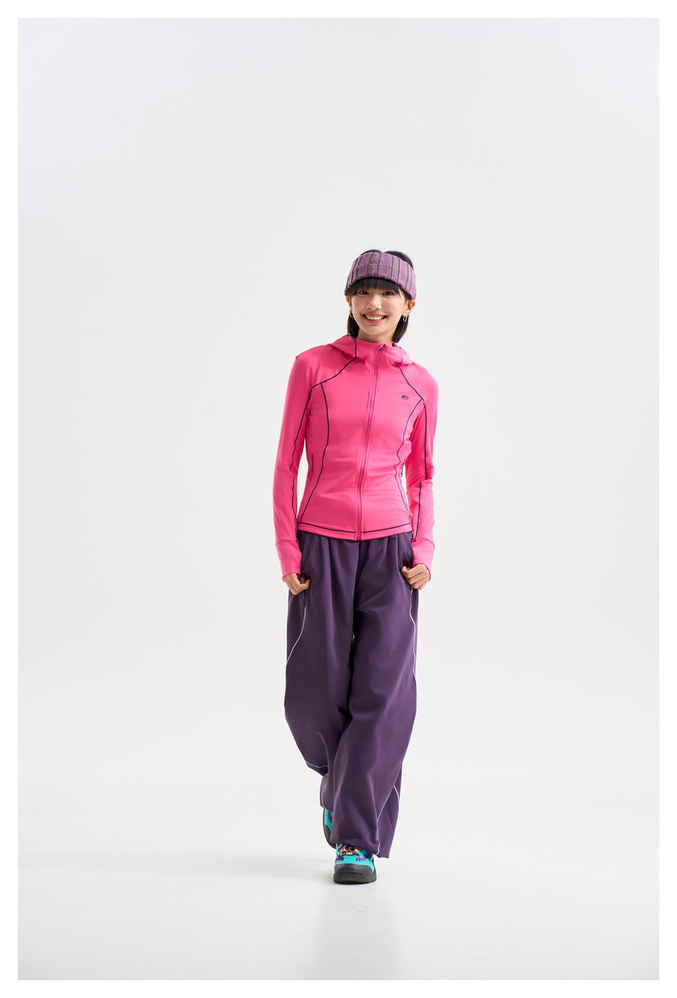 Model wearing a bright pink zip-front hooded jacket with dark piping and loose purple pants, walking toward the camera.