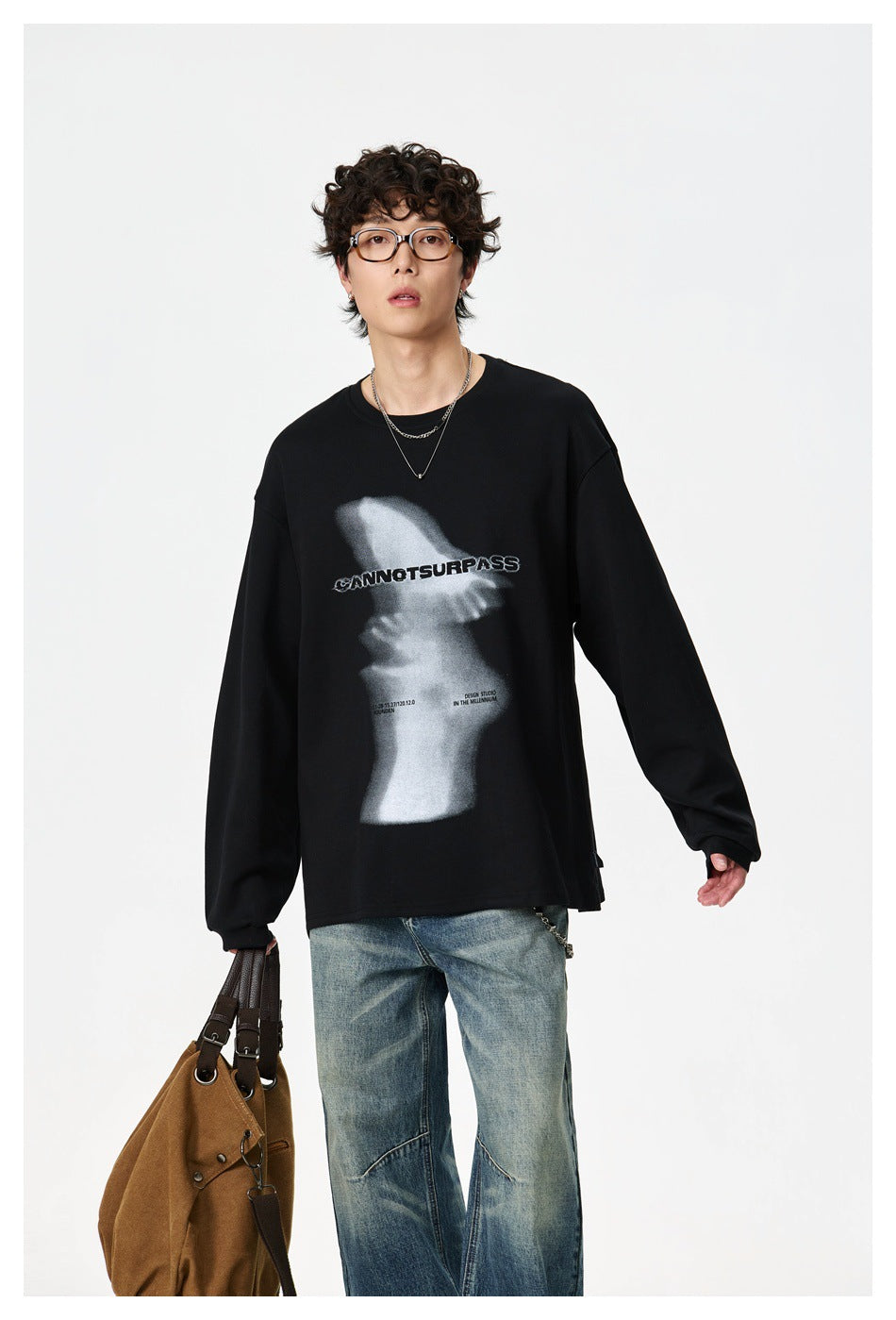 Model wearing a black sweatshirt with a large grayscale front graphic and loose blue jeans, carrying a brown bag.