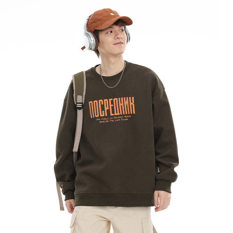 Model wearing a dark olive crewneck sweatshirt with orange chest lettering and ribbed cuffs.
