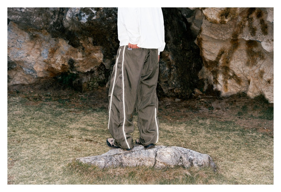 Rear view of a model wearing loose taupe-gray pants with reflective piping along the legs.