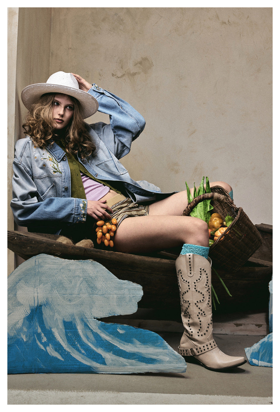 Model seated in an oversized light blue embroidered jacket, striped shorts, white hat, and beige boots.