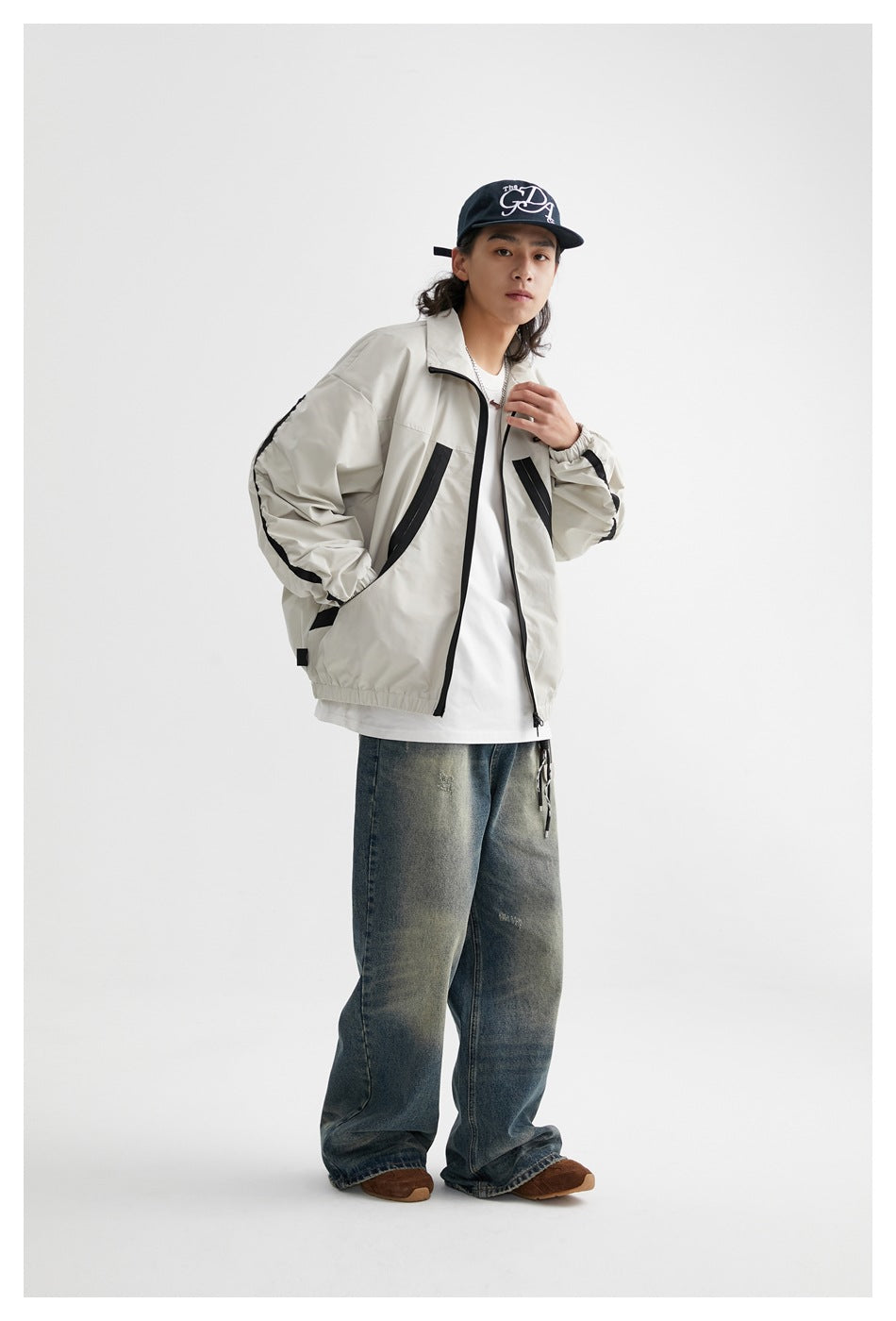 Full-length model wearing a light gray zip jacket with black trim, loose faded jeans, and a dark cap.