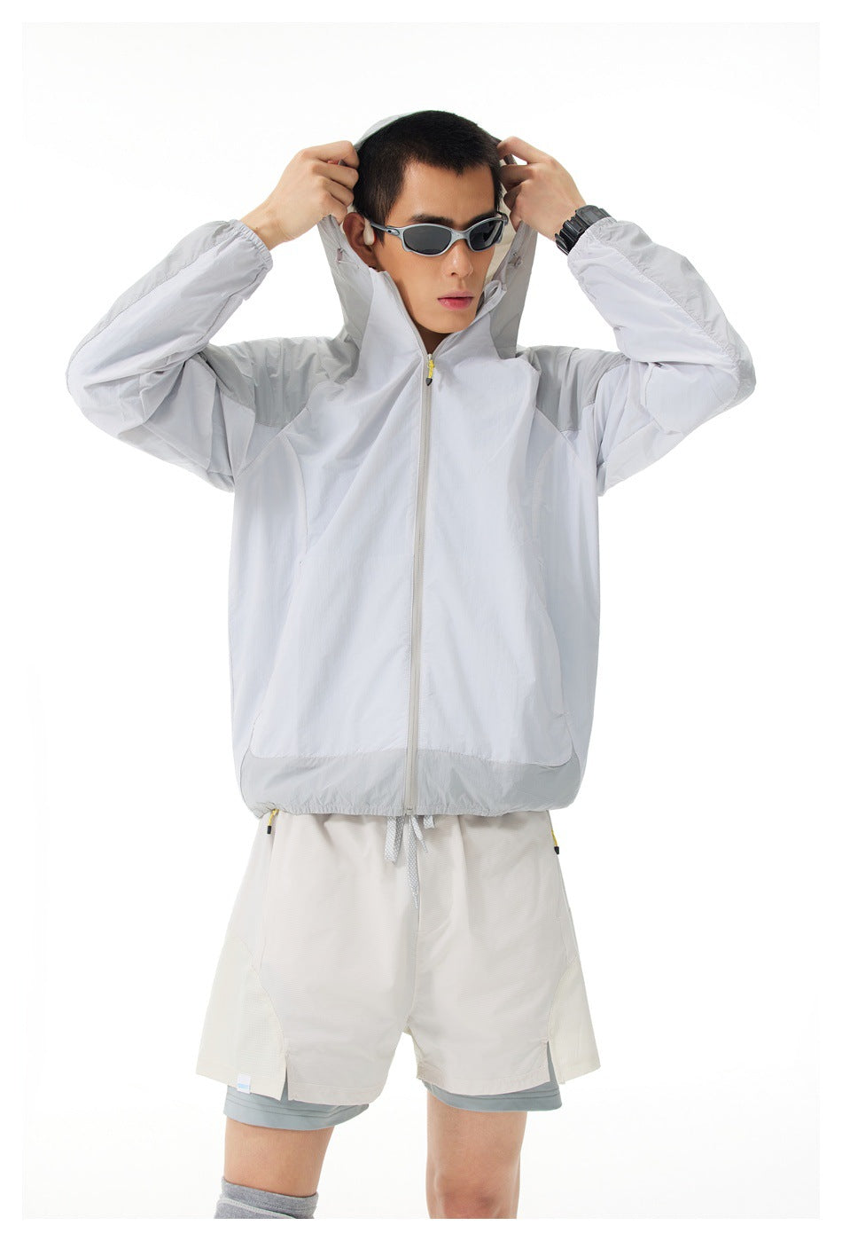 Closer view of a model lifting the hood of a light gray zip jacket, worn with layered white shorts.