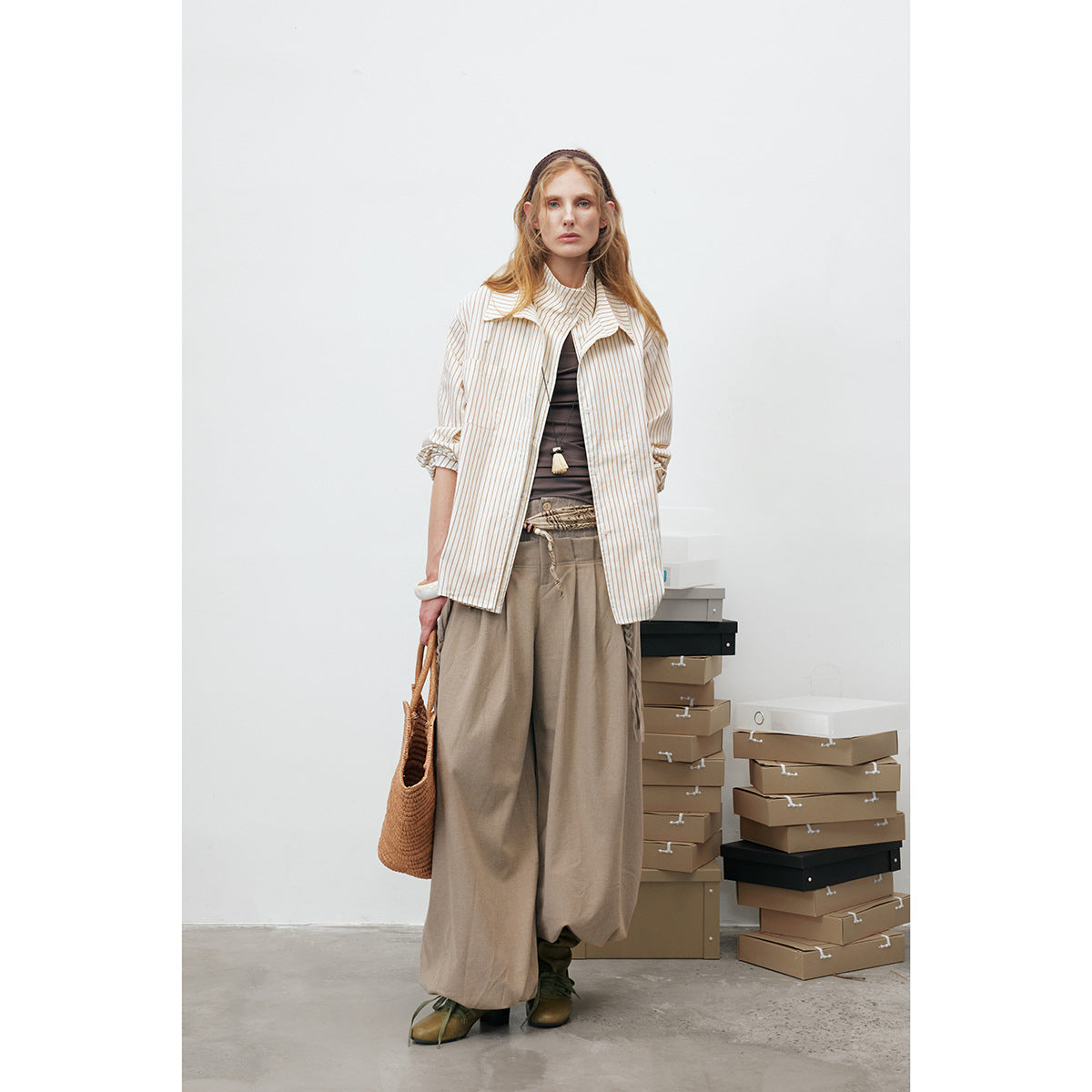 Model wearing an ivory shirt with thin tan stripes over wide taupe trousers, carrying a woven bag.