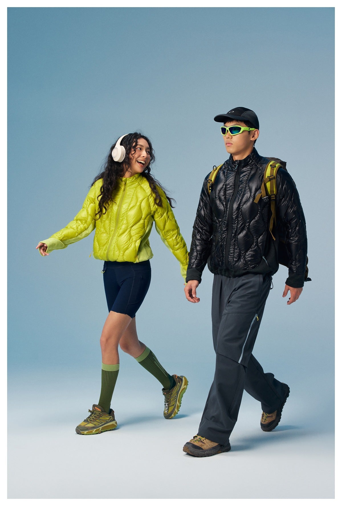 Two models walk in lime and black quilted jackets, with hiking gear and a blue studio backdrop.