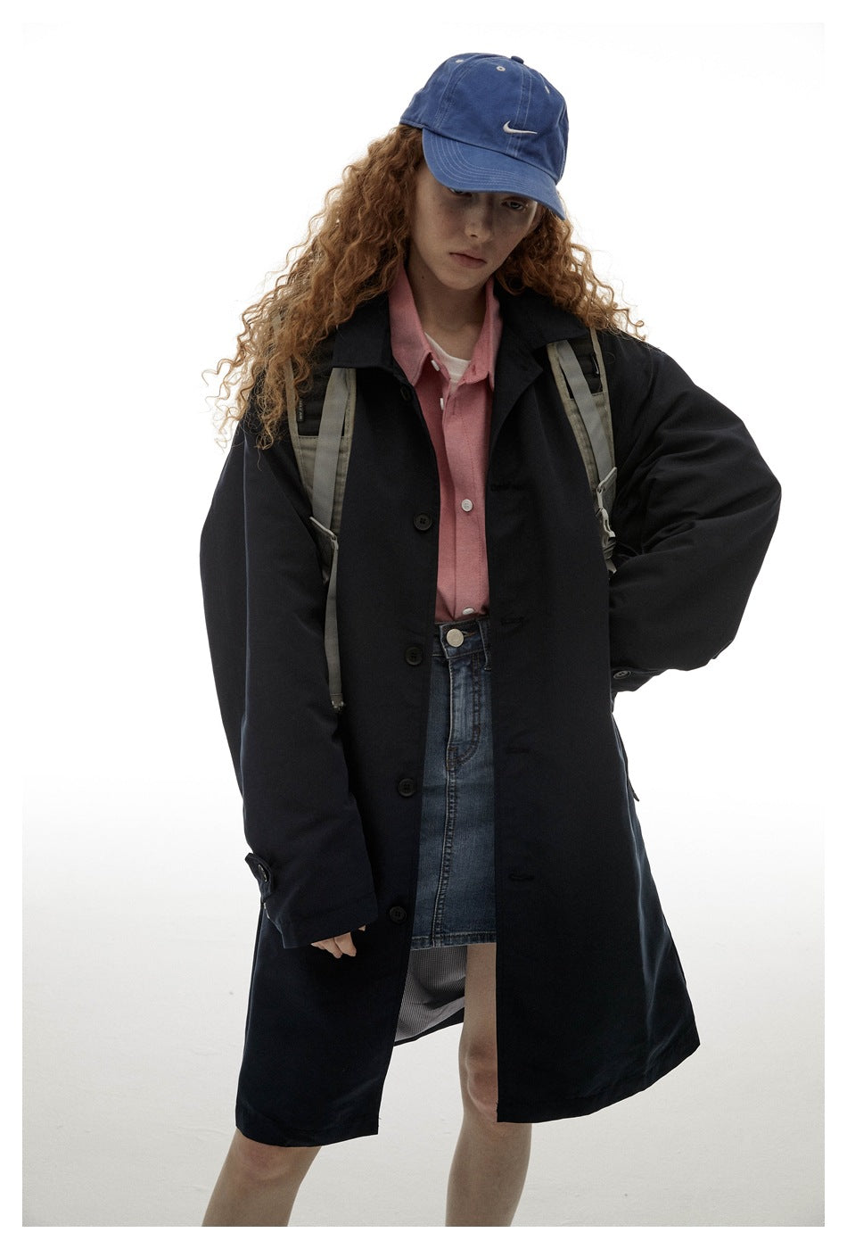 Model wearing a dark navy button-front coat over a pink shirt and denim skirt, with a blue cap and backpack.