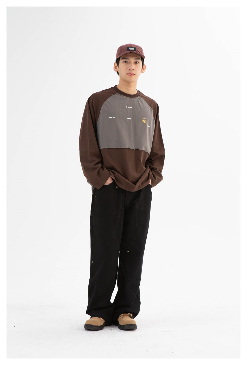 Full-length model wearing a brown and gray color-block long-sleeve shirt with black pants and a burgundy cap.