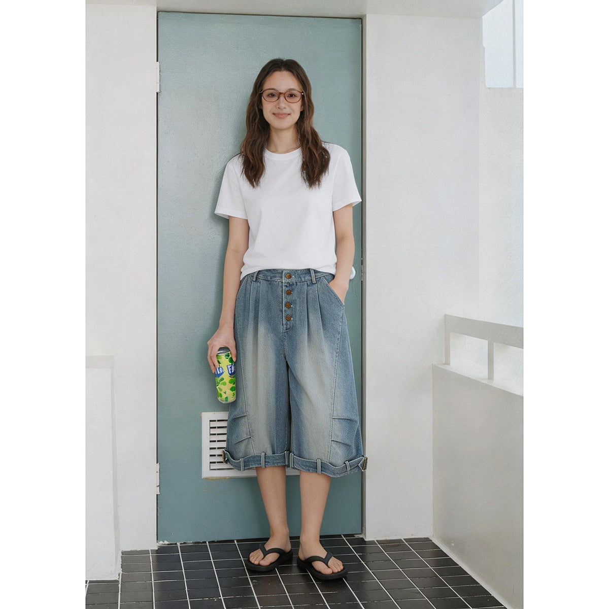 Woman wearing faded blue knee-length wide-leg denim shorts with a white T-shirt and black sandals, standing by a door