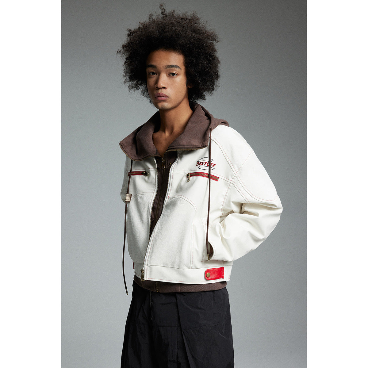 Model wearing a cropped white jacket with red zip details over a brown hooded layer and black trousers.