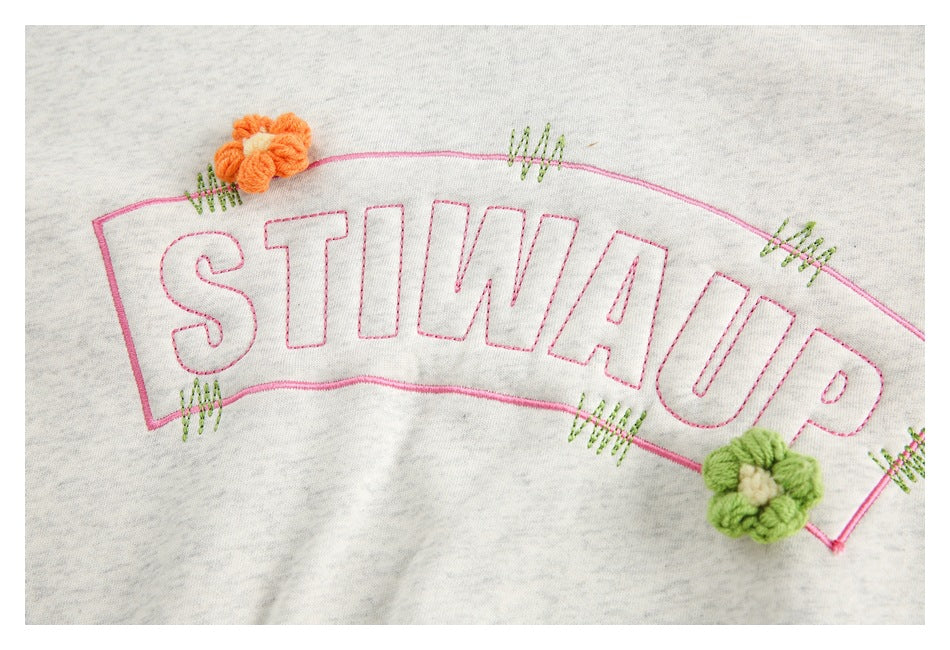 Close-up of outlined pink lettering with small orange and green flower embroidery on light gray heather fabric.