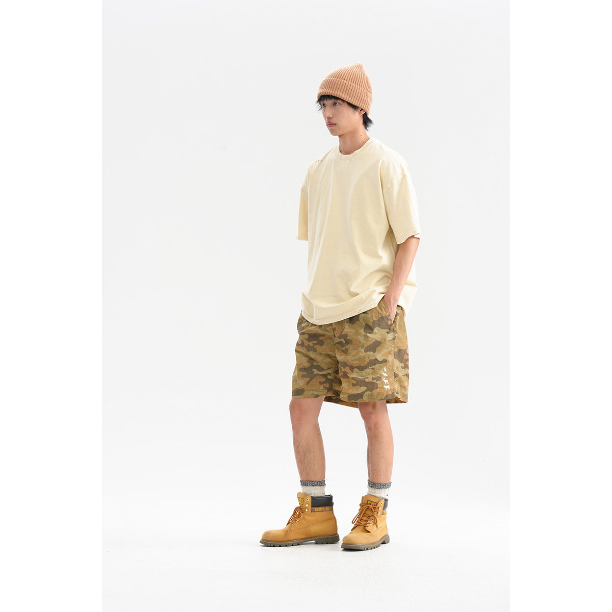 Man wearing an oversized cream T-shirt and tan camouflage shorts, shown in a three-quarter view against a white background.