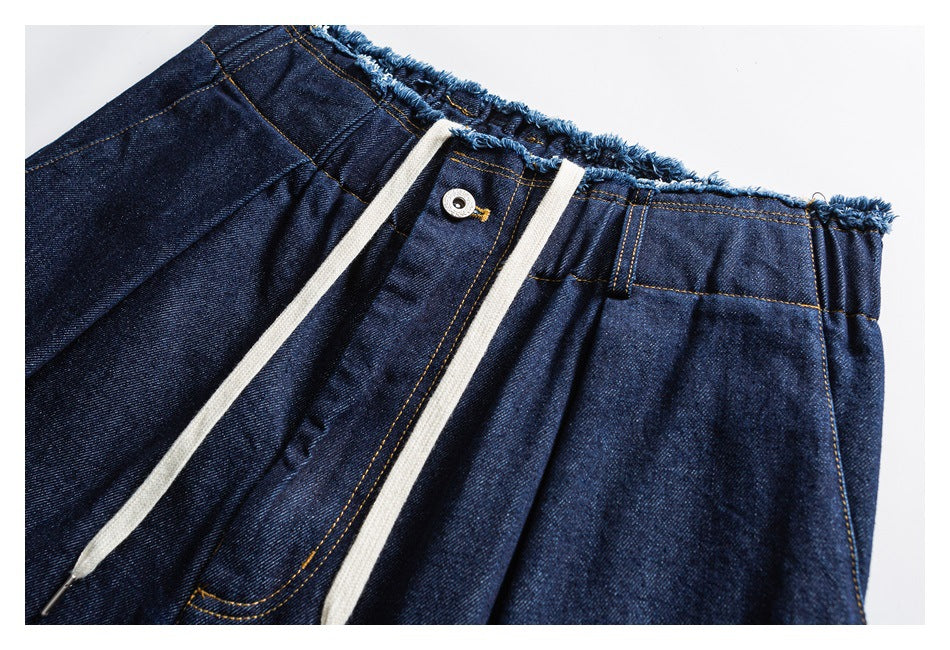 Close-up of dark indigo denim pants with a frayed waistband, button fly, and cream drawstrings.