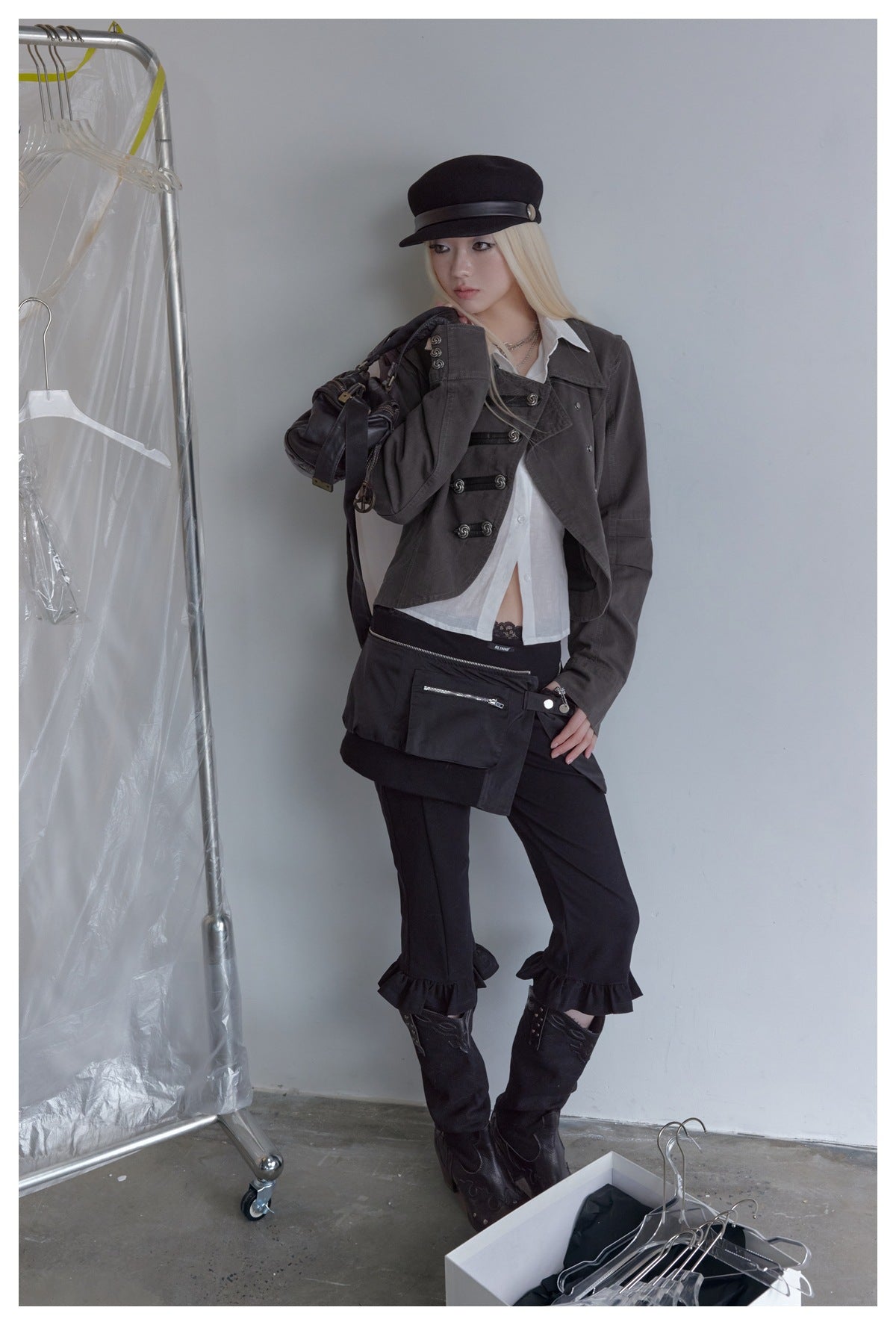 Model in a dark gray cropped double-breasted jacket with white shirt, black cap and holding a bag, leaning on a wall