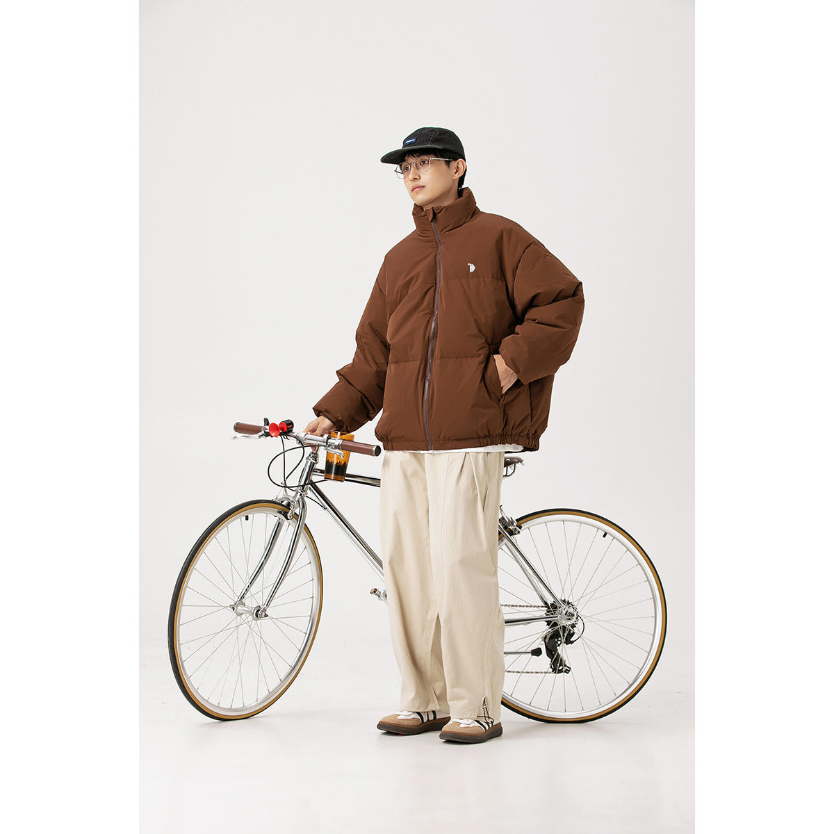Model wearing a brown high-collar puffer jacket beside a bicycle in a studio.