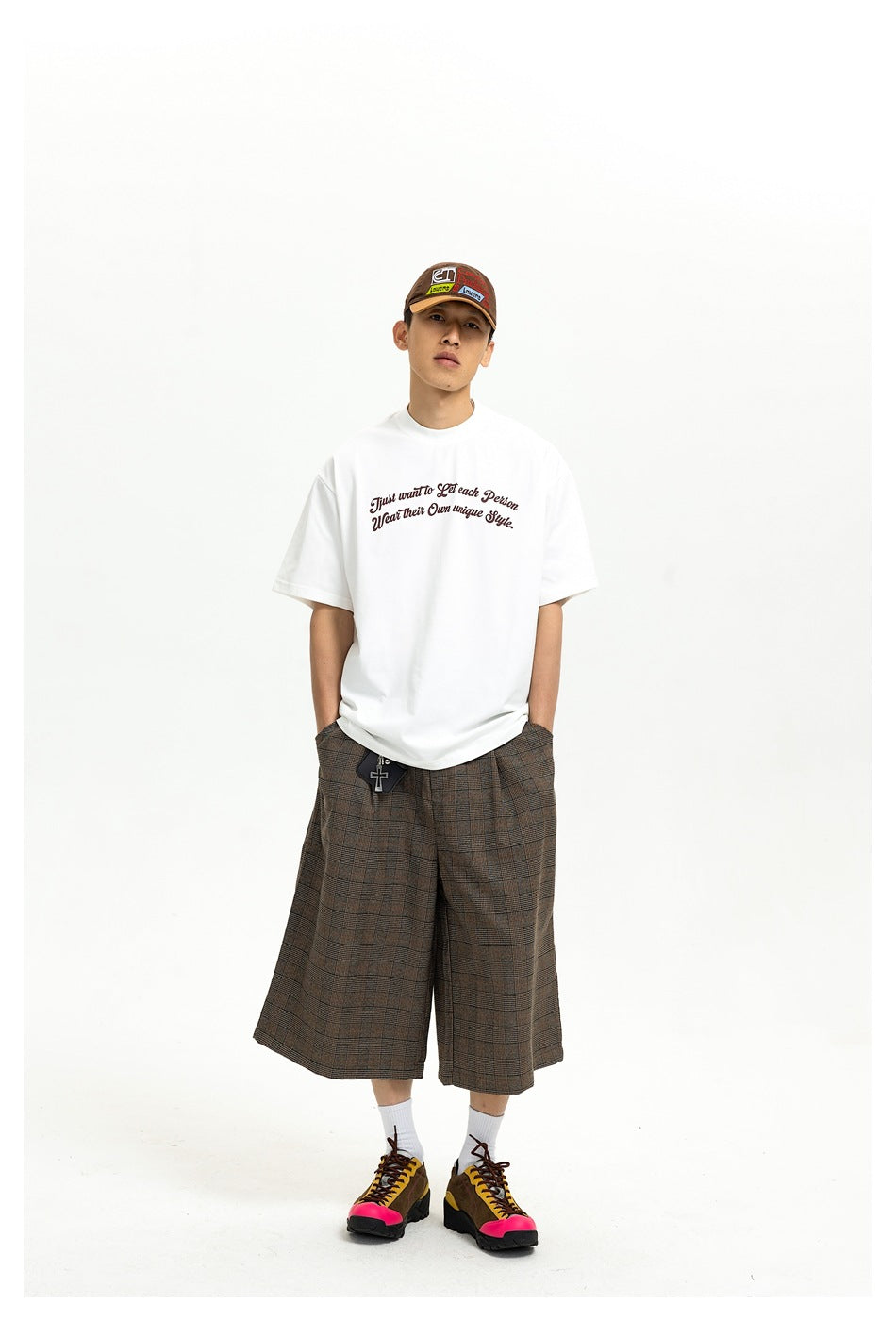 Man wearing a white slogan T-shirt with brown plaid shorts, a cap and colorful sneakers, standing front view