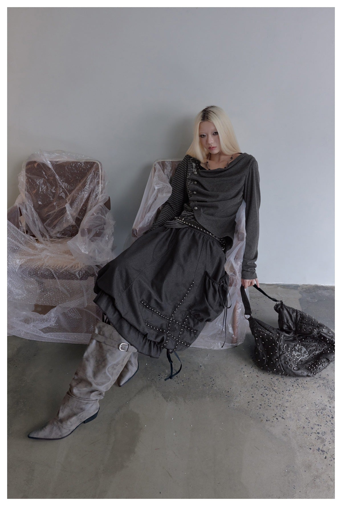 Seated model wearing a black bubble skirt with a studded cross patch and ribbon ties, with gray boots and a studded bag