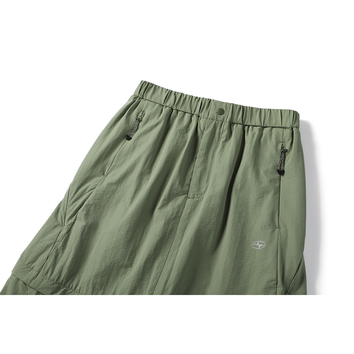 Cropped close-up of an olive-green skirt front showing an elastic waist, snap closure, zip pockets, and small emblem.