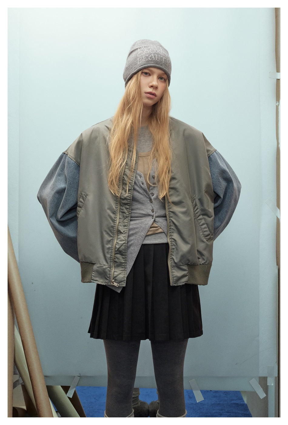 Front view of a model wearing an oversized olive-gray jacket with blue denim sleeves and ribbed cuffs over a black pleated