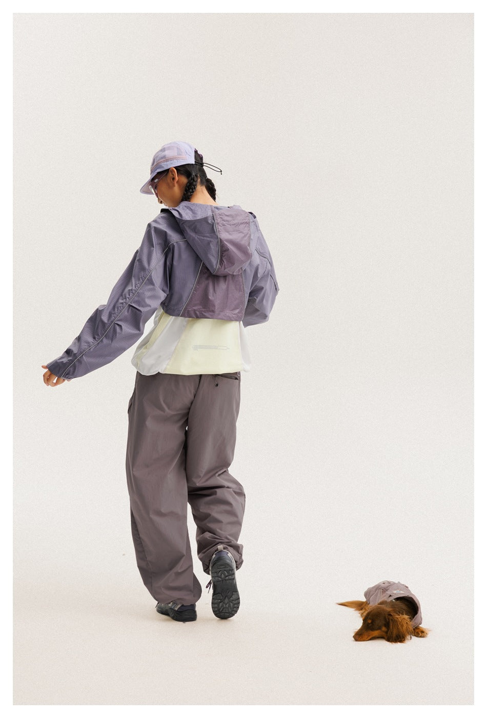Rear view of a model wearing a purple hooded windbreaker over a pale top and loose gray-purple pants.