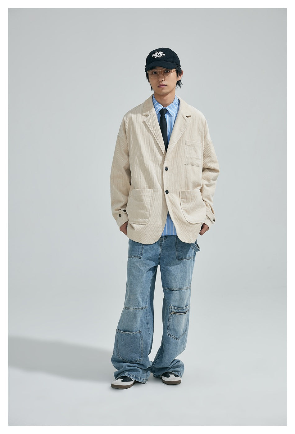 Full-length front view of a model wearing a cream corduroy blazer, blue striped shirt, black tie, and wide-leg jeans.
