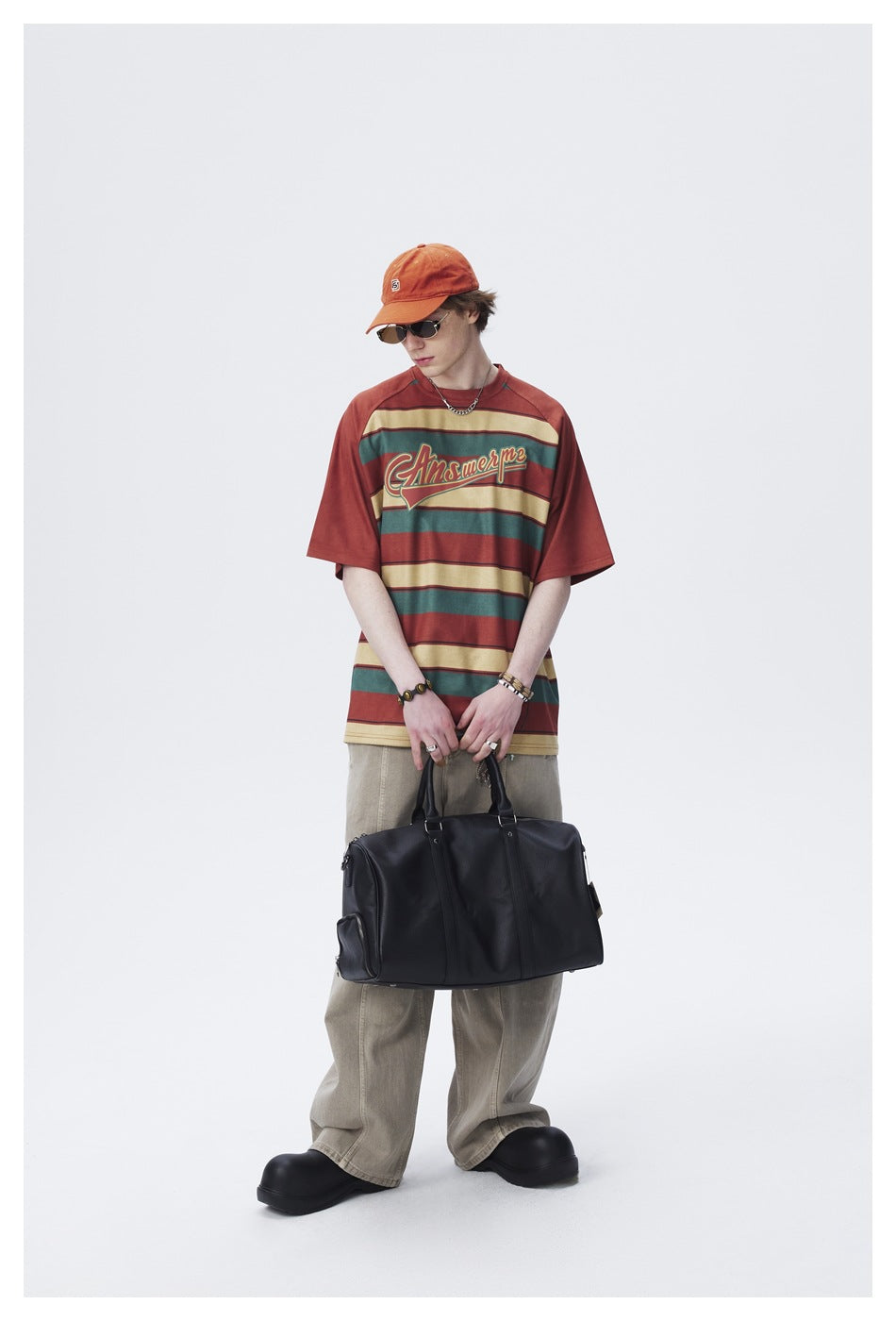 Full-length model holding a black bag and wearing a rust, teal and cream striped graphic T-shirt.