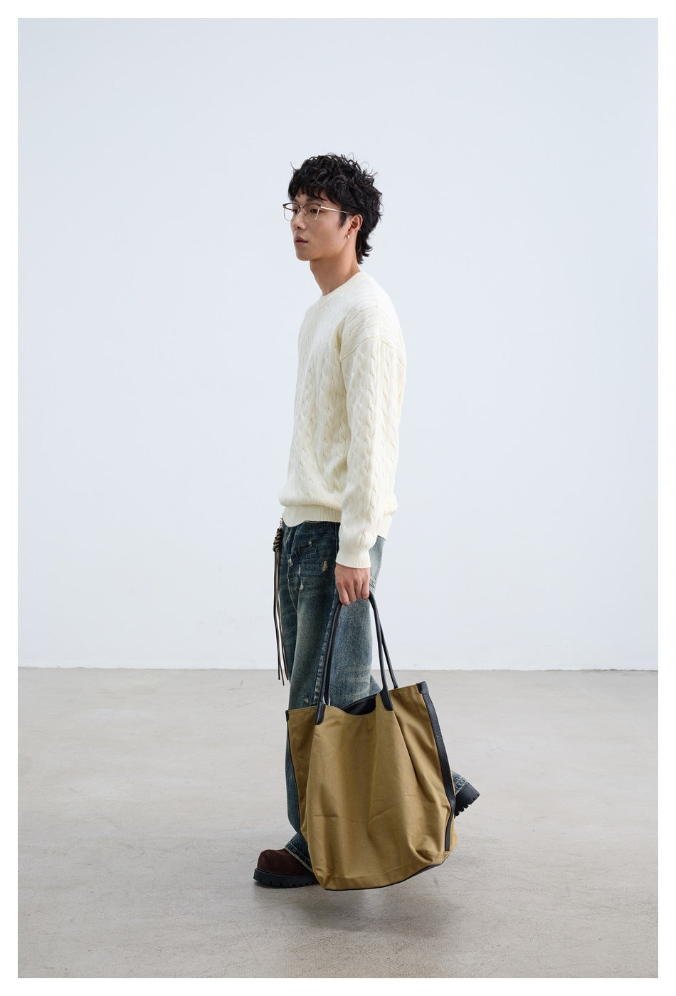 Side-profile view of a model wearing an ivory cable-knit crewneck sweater and dark jeans, holding a tan tote.