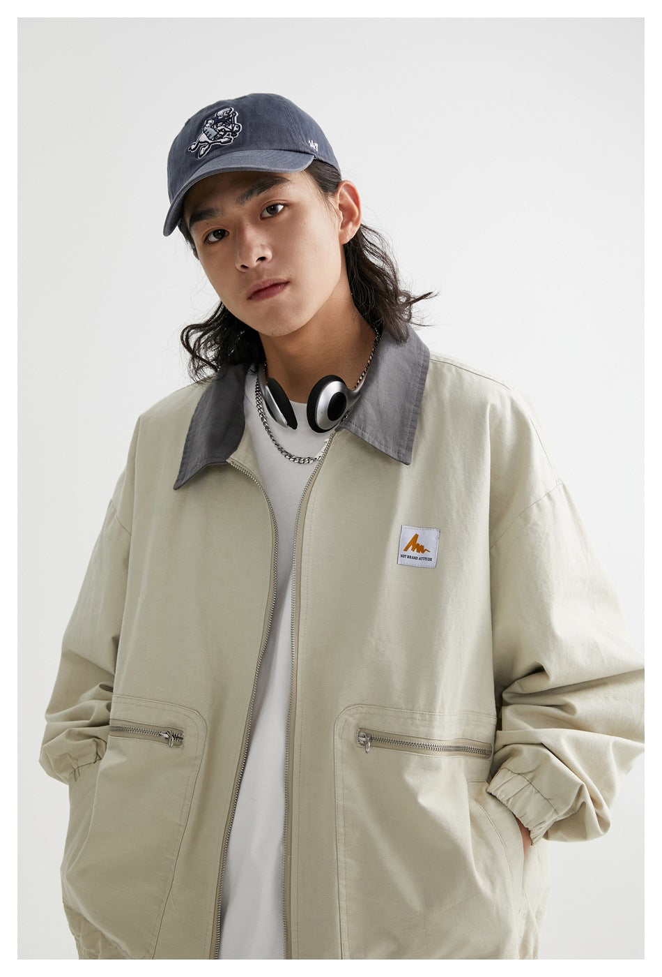 Close view of a model wearing a pale beige jacket with a gray collar, front zipper, elastic cuffs, and zip pockets.