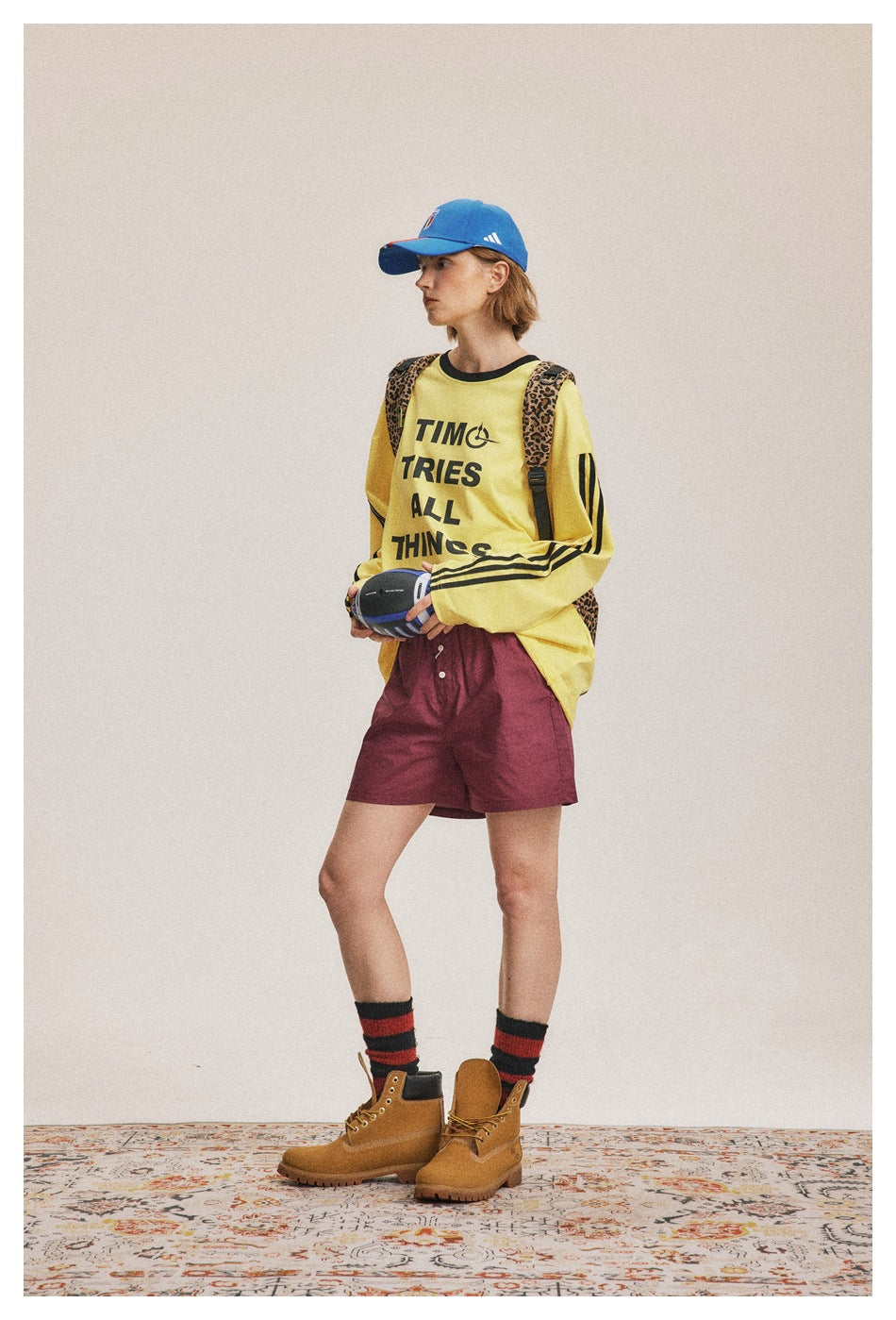 Model in side pose wearing the yellow lettered long-sleeve top with burgundy shorts, blue cap and leopard backpack