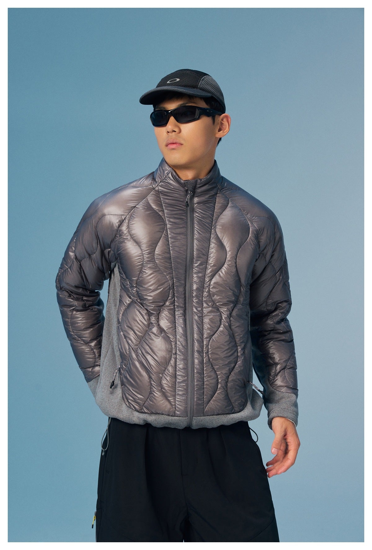 Model wearing a shiny charcoal quilted jacket with a high collar, gray knit side panels and cuffs, and a front zip.