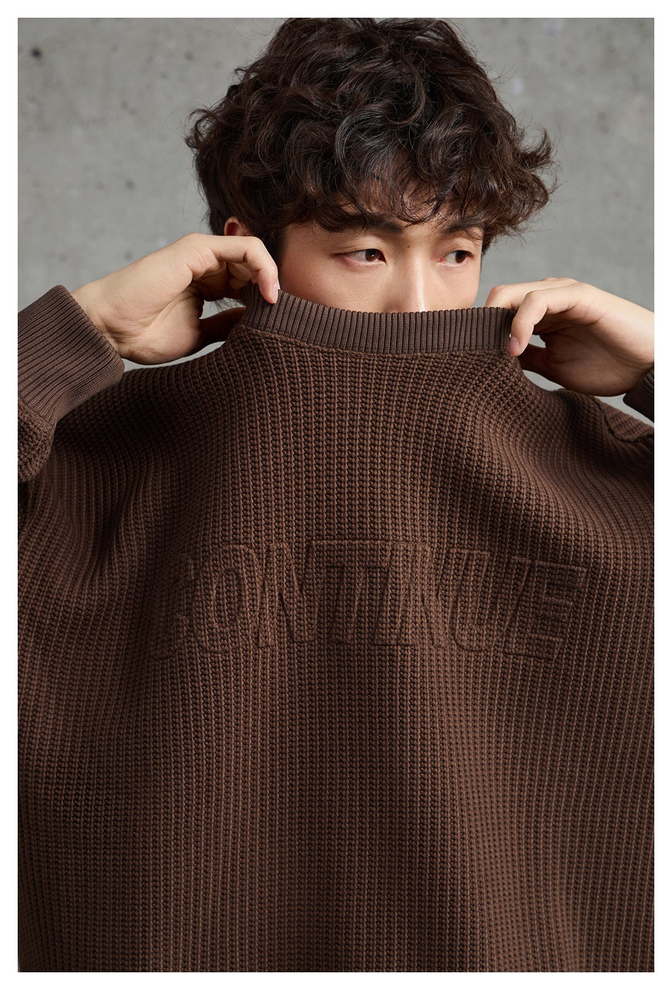 Close-up of a model holding up a dark brown textured crewneck sweater to show its ribbed collar and tonal chest lettering.