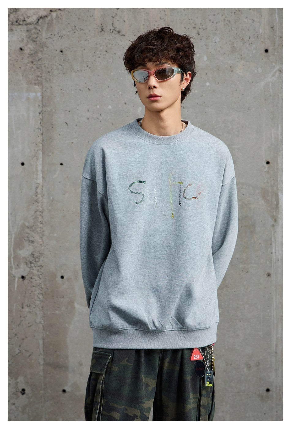 Model wearing a light gray crewneck sweatshirt with subtle multicolored chest lettering and dark camouflage cargo pants.