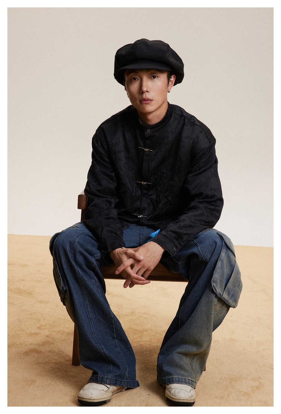 Seated model wearing a black textured stand-collar jacket with frog closures and tonal embroidery.