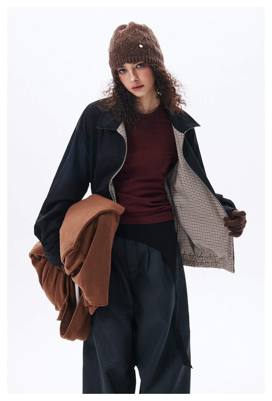 Model holds open a black jacket to show its checked lining, worn over a burgundy top with a brown knit beanie.