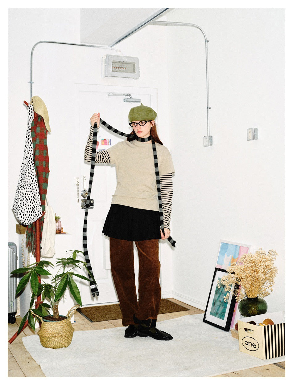 Front view of a model in a beige layered top, black skirt, brown trousers, green beret and long black-and-gray striped scarf.