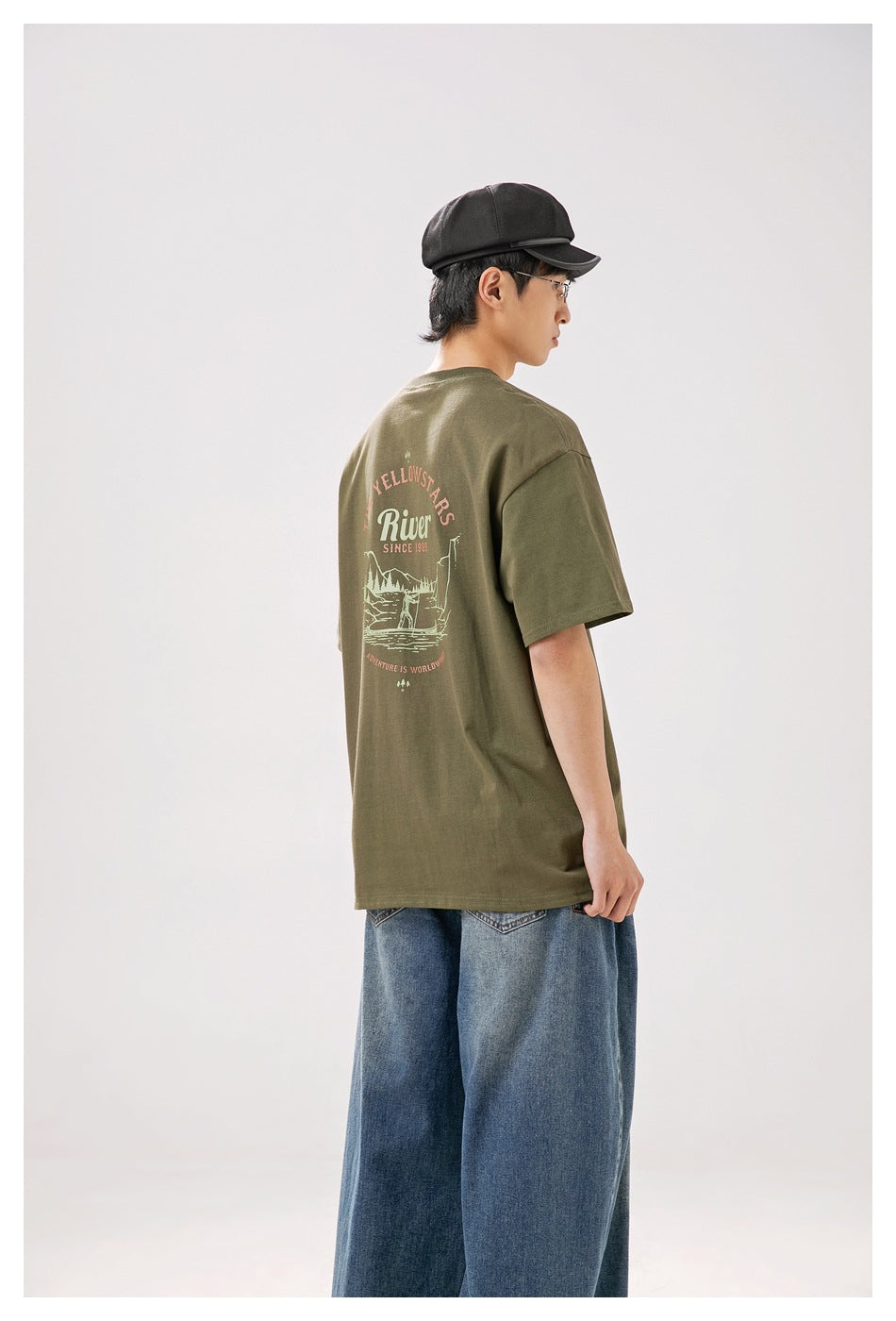 Rear three-quarter view of a model wearing an olive T-shirt with a large outdoor-themed graphic on the back.