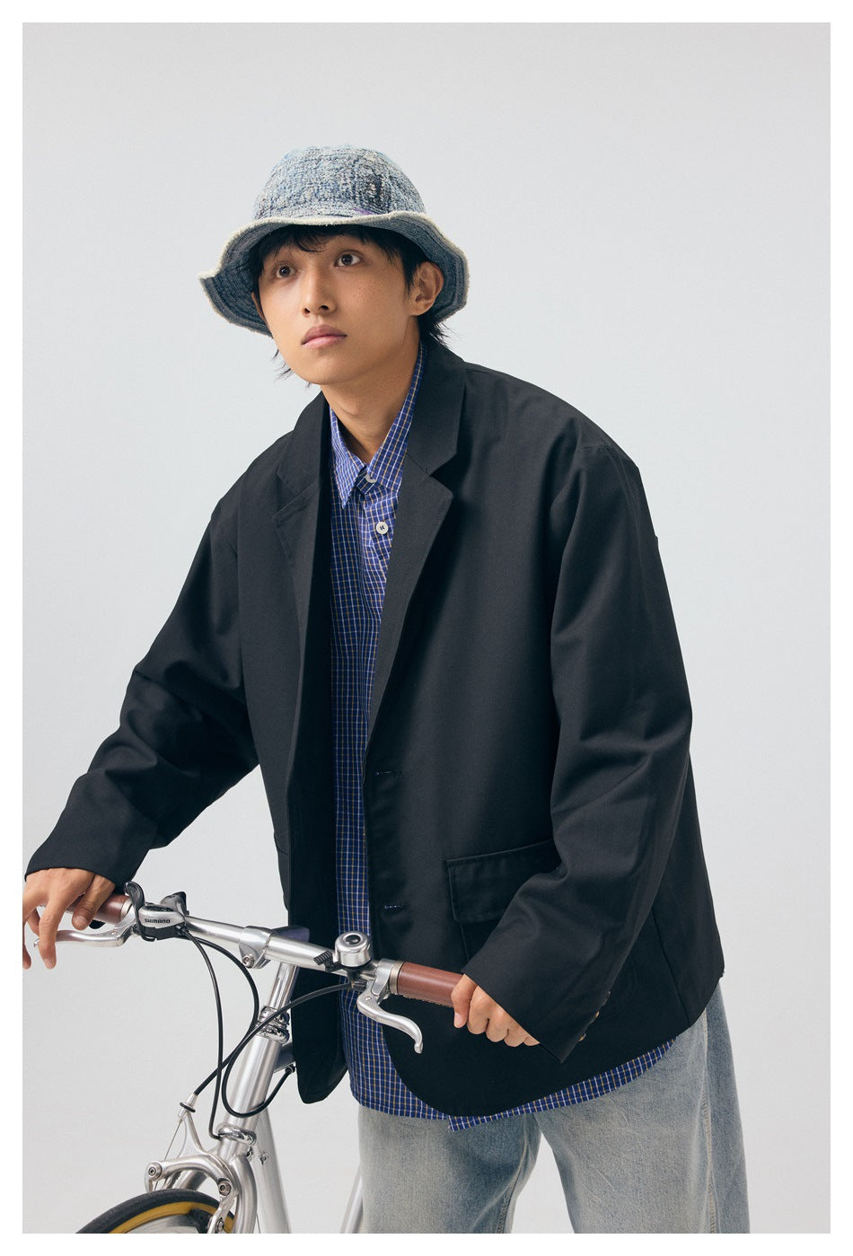 A person models an oversized black blazer over a blue checked shirt, shown leaning on a bicycle.