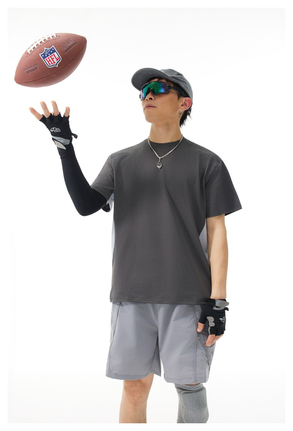 Model in a dark gray shirt and light gray shorts tosses a football, wearing gloves, a cap, and sunglasses.