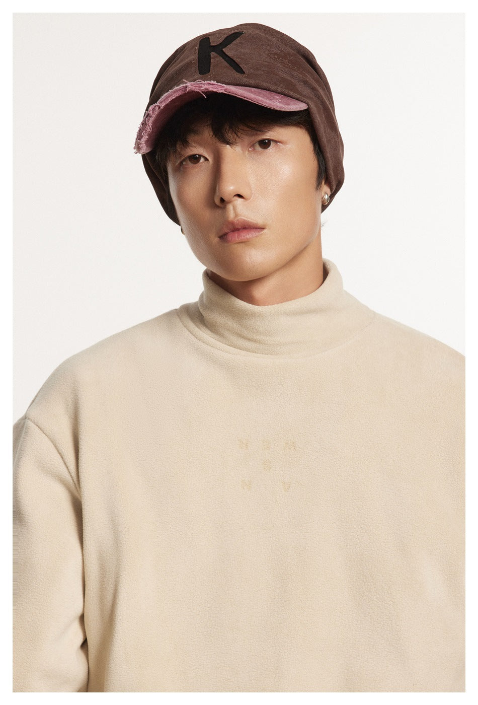 Model wearing a cream high-neck fleece pullover and a brown cap with a distressed pink brim.