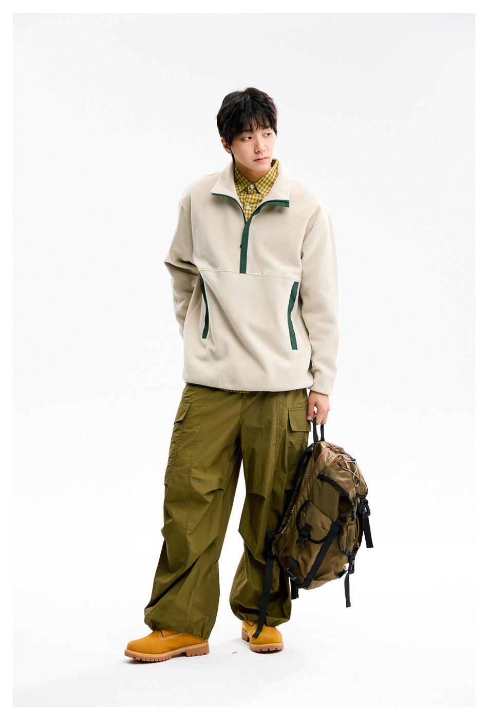 Model in beige half-zip pullover with green trim and olive cargo pants, looking sideways while holding a backpack